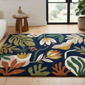 Puede incluir: Una alfombra rectangular con un fondo azul marino profundo y un patrón botánico a gran escala. El diseño incluye hojas y flores estilizadas en tonos verde oliva, naranja quemado, crema y amarillo mostaza en toda la superficie.