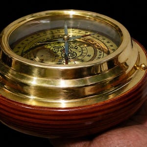 May include: A vintage brass compass with a wooden base. The compass has a clear glass top, a gold-coloured dial, and a needle pointing north. The wooden base is a rich, dark brown colour. The compass is held in a hand.