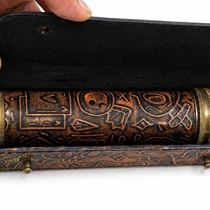 May include: A cylindrical, brown, embossed leather object with brass accents, housed in a matching leather case. The object features intricate patterns and symbols. The case is open, revealing the contents. The overall design suggests a vintage or antique aesthetic.