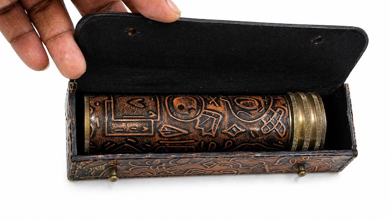 May include: A cylindrical, brown, embossed leather object with brass accents, housed in a matching leather case. The object features intricate patterns and symbols. The case is open, revealing the contents. The overall design suggests a vintage or antique aesthetic.