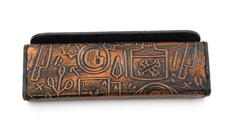 May include: A brown leather case with a black top, featuring embossed designs of knives, poison bottles, and other symbols. The case has a distressed, vintage look, with a dark brown and black color scheme.