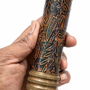 May include: A cylindrical, decorative object with a copper and dark green finish. The object has an embossed pattern and is stitched with brown thread. The base and top are brass-colored. The object is held in a hand.