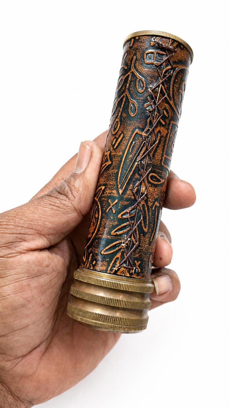 May include: A cylindrical, decorative object with a copper and dark green finish. The object has an embossed pattern and is stitched with brown thread. The base and top are brass-colored. The object is held in a hand.