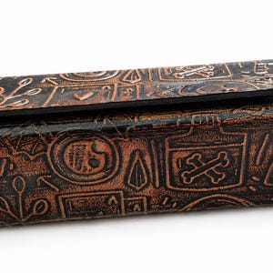 May include: A rectangular, brown leather box with a hinged lid. The box is embossed with various symbols, including skulls, crossbones, and botanical elements. The leather has a distressed, antique appearance.