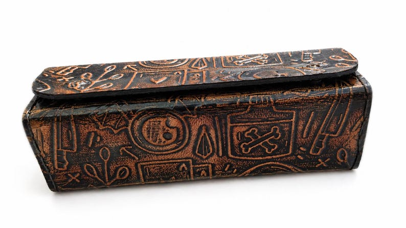 May include: A rectangular, brown leather box with a hinged lid. The box is embossed with various symbols, including skulls, crossbones, and botanical elements. The leather has a distressed, antique appearance.