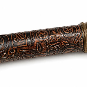 May include: A cylindrical, antique-style object with a dark brown, embossed leather exterior. The design features skulls, crossbones, and other intricate patterns. The ends are capped with brass-colored metal. The object is likely a container or decorative item.