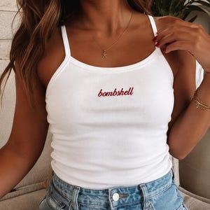 May include: A white ribbed tank top with thin shoulder straps featuring the word bombshell embroidered in red cursive script across the center of the chest. The garment is paired with light blue denim jeans with a visible silver metal button.