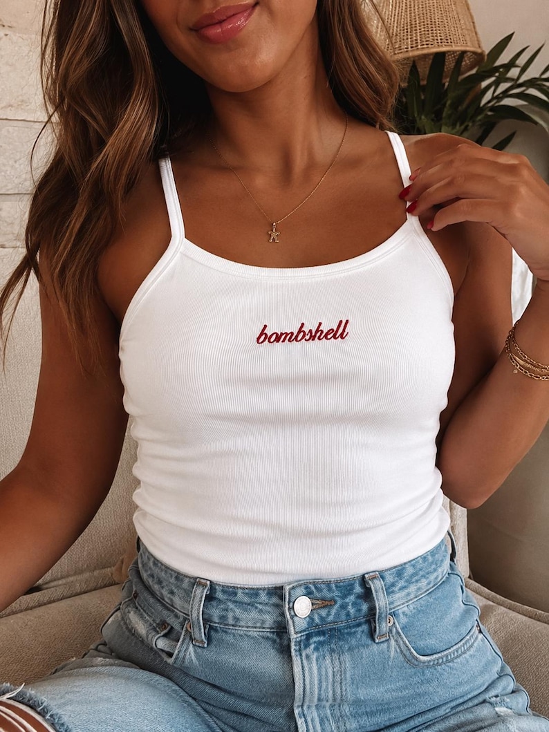 May include: A white ribbed tank top with thin shoulder straps featuring the word bombshell embroidered in red cursive script across the center of the chest. The garment is paired with light blue denim jeans with a visible silver metal button.