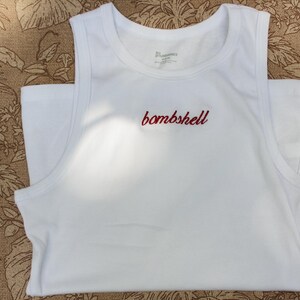 May include: A white sleeveless tank top featuring a crew neckline and wide shoulder straps. The word bombshell is embroidered in red cursive script across the center of the chest. The garment is made of a solid white fabric with a smooth texture.