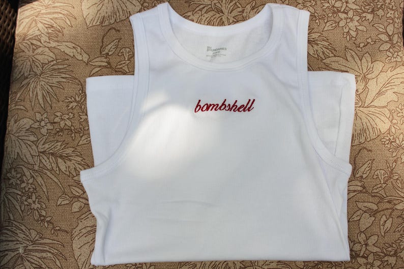 May include: A white sleeveless tank top featuring a crew neckline and wide shoulder straps. The word bombshell is embroidered in red cursive script across the center of the chest. The garment is made of a solid white fabric with a smooth texture.