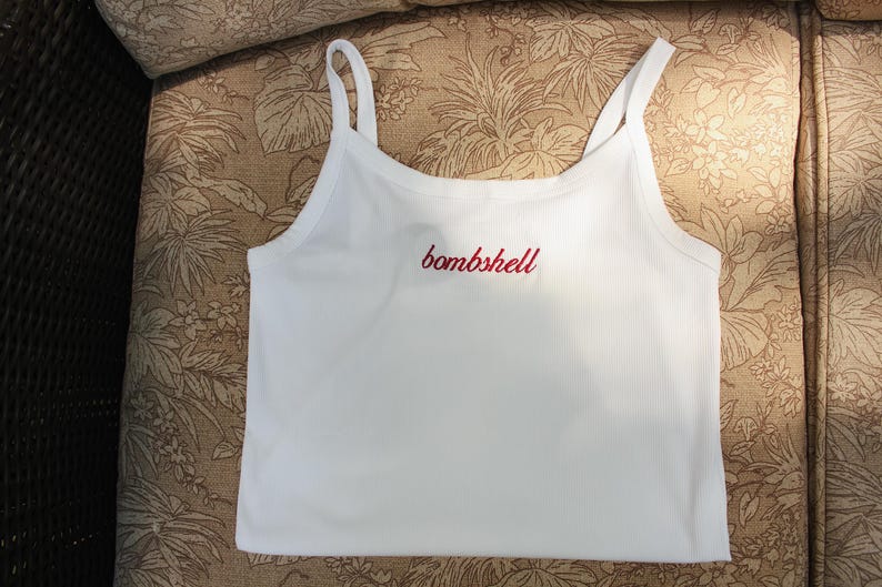 May include: A white ribbed tank top with thin shoulder straps featuring the word bombshell embroidered in red cursive script across the center of the chest. The garment is constructed from a textured fabric and has a straight hemline.