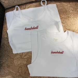 May include: Two white sleeveless tops made of fabric. One top features thin spaghetti straps and a scoop neckline, while the other is a tank top style with wider shoulder straps. Both garments have the word bombshell embroidered in red script.