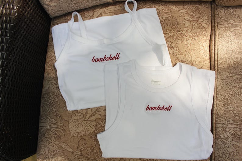 May include: Two white sleeveless tops made of fabric. One top features thin spaghetti straps and a scoop neckline, while the other is a tank top style with wider shoulder straps. Both garments have the word bombshell embroidered in red script.