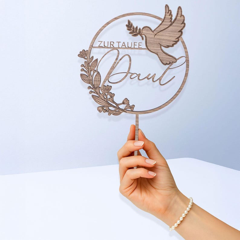 Eine Hand hält den personalisierten Cake Topper zur Taufe und zeigt seine Größe im Verhältnis zur Hand. Runder Tortenaufsatz mit Taube, Namen und floralen Zweigen.