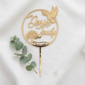 Goldfarbener Cake Topper „Zur Taufe“ mit rundem Rahmen, Taubenmotiv und filigranen Blumen auf hellem Stoff neben einem Eukalyptuszweig.