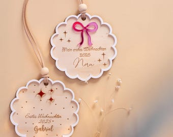 May include: Two wooden Christmas ornaments with scalloped edges and a light wood grain. One features a pink bow and the text "Mein erstes Weihnachten 2025 Nora." The other has the text "Erstes Weihnachten 2025 Gabriel." Both have a string for hanging.