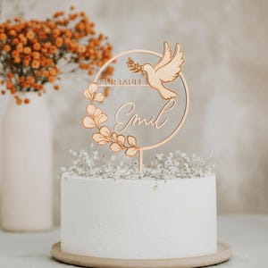 Goldfarbener Cake Topper mit Namen, Taube und Eukalyptuskranz auf einer weißen Torte mit zarten Blüten vor einem Hintergrund mit Trockenblumen.