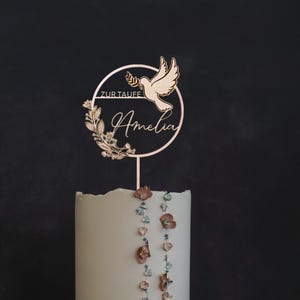 Può includere: Un cake topper in legno con una colomba che porta un ramoscello d'ulivo, una ghirlanda floreale e il nome "Amelia". Le parole "Zur Taufe" sono sopra il nome. Il topper è su una torta bianca con perline decorative.