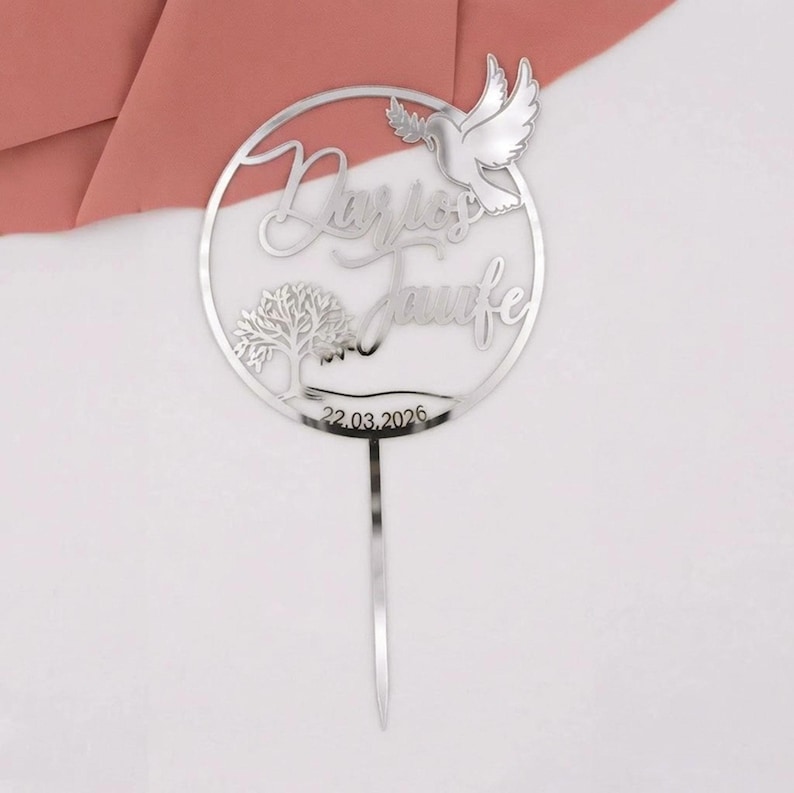 Silberner Tortentopper „Zur Taufe“ mit rundem Rahmen, geschwungener Schrift, Taube und Blumenmotiv auf weißem Hintergrund mit rosafarbenem Stoff.