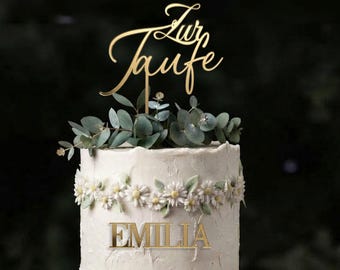 May include: A white frosted cake decorated with a horizontal band of white daisy flowers and a gold acrylic name plaque reading EMILIA. A gold cake topper with the German script text Zur Taufe sits above green eucalyptus foliage.