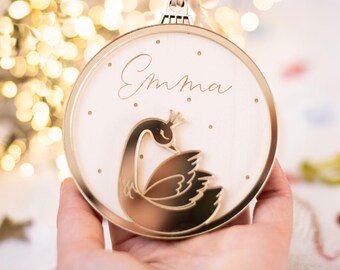 May include: A round Christmas ornament with a gold-colored frame and a white interior. The ornament features a gold swan design and the name "Emma" in cursive. Small gold dots are scattered around the name. A beige ribbon is attached for hanging.