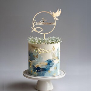 Kommuniontorte mit goldenem Cake Topper „Zur Erstkommunion“, Taube und Fischsymbol sowie personalisiertem Namen „Melanie“ und weißen Blüten.