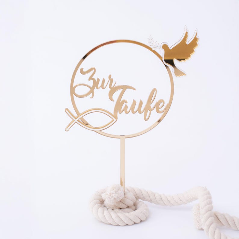 Ein goldener Tortentopper mit der Aufschrift Zur Taufe steckt in einem eingerollten beigen Seil auf weißem Grund.