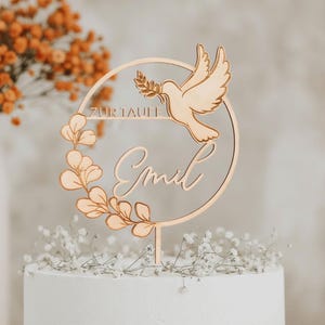 Personalisierter Cake Topper aus goldfarbenem Spiegelacryl mit Taube, Eukalyptuskranz und Namensschriftzug auf einer weißen Torte vor orangefarbenen Trockenblumen.