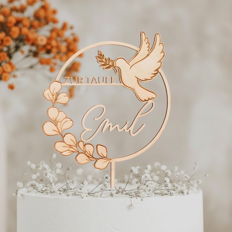Personalisierter Cake Topper aus goldfarbenem Spiegelacryl mit Taube, Eukalyptuskranz und Namensschriftzug auf einer weißen Torte vor orangefarbenen Trockenblumen.