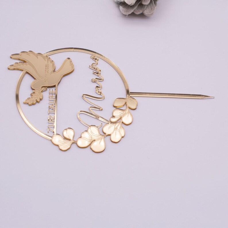 Personalisierter goldfarbener Tortenstecker mit Taube, rundem Rahmen und Eukalyptusblättern, flach auf einem hellen Hintergrund fotografiert.