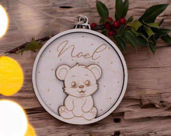 May include: Wooden Christmas ornament with a bear design. The ornament is round, with the word "Noel" in gold script. A cute bear is in the center. The ornament is made of light-colored wood and has a string for hanging.