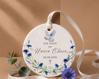 May include: A round white ceramic ornament featuring a blue dove illustration, a floral wreath of blue cornflowers, and the text ZUR TAUFE von Hanna Olivia 28.09.2026 with a small blue cross. A white satin ribbon is attached for hanging.