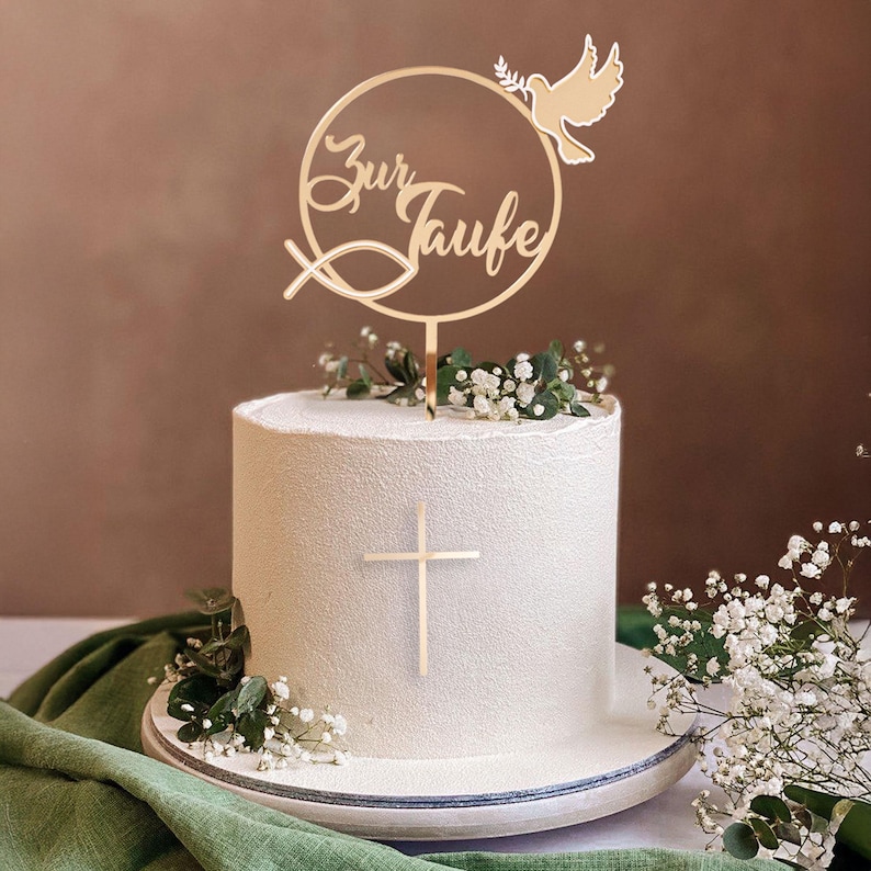 Ein runder goldener Tortenstecker mit der Aufschrift Zur Taufe, einer Taube und einem Fisch steckt in einer weißen Torte mit goldenem Kreuz und Eukalyptus.