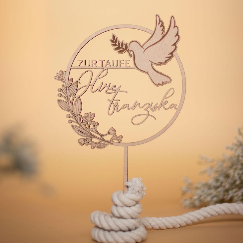Personalisierter Cake Topper „Zur Taufe“ mit filigranem Namensschriftzug, Taube und Blumenranke, auf einem weißen Knotenhalter vor beigem Hintergrund.