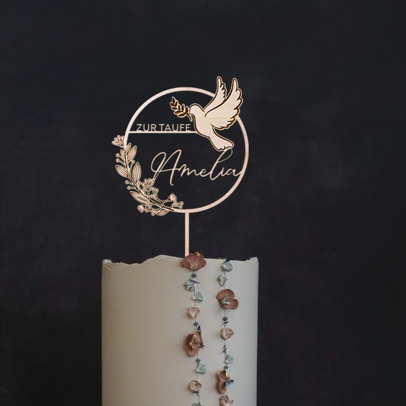 Personalisierter Cake Topper zur Taufe mit Namen Amelia, Taube und floralen Zweigen im runden Rahmen auf einer weißen Tauftorte vor dunklem Hintergrund.