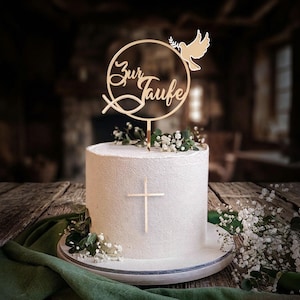 Eine weiße Torte mit goldenem Kreuz und dem runden Tortentopper Zur Taufe, dekoriert mit Schleierkraut.