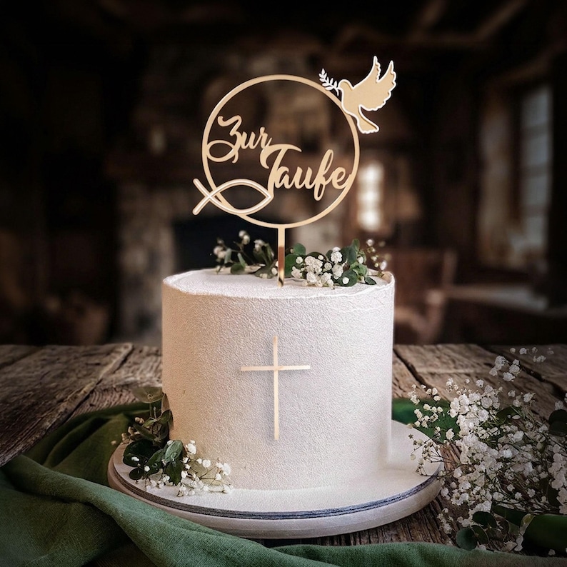 Eine weiße Torte mit goldenem Kreuz und dem runden Tortentopper Zur Taufe, dekoriert mit Schleierkraut.