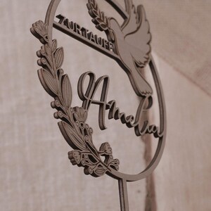Nahaufnahme des runden Cake Toppers zur Taufe mit personalisiertem Namensschriftzug, Taube und filigranen Blätterranken vor hellem Stoff.
