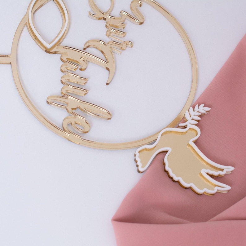 Nahaufnahme des goldenen runden Tortentoppers mit Taube und der Aufschrift Zur Taufe auf rosa Stoff.