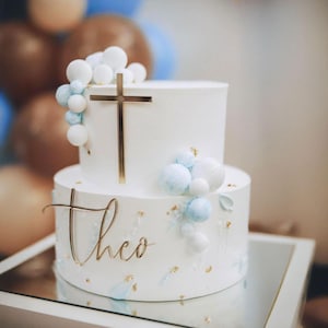 May include: Two-tiered white cake decorated with gold cross and the name "Theo". The cake is adorned with white and light blue spherical decorations and gold accents. The cake sits on a mirrored surface, with blurred balloons in the background.