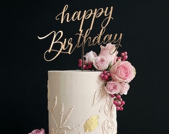 May include: A white frosted cake decorated with pink roses, berries, and gold leaf accents. A gold "Happy Birthday" cake topper is placed on top. The cake is set against a black background.