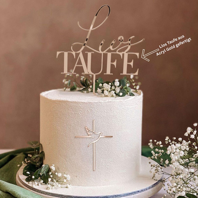 May include: A white frosted cake with a gold-colored topper that reads "Liz Taufe". The cake is decorated with a gold cross and dove design, and fresh white flowers. The text "Lios Taufe aus Acryl Gold gefertigt" is also visible.