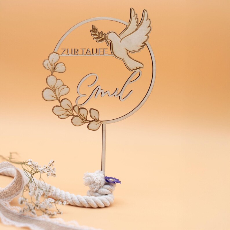 Cake topper taufe acryl in Gold mit Namen „Paul“, rundem Eukalyptuskranz und Taube, auf einem Stiel vor warmem beigefarbenem Hintergrund.