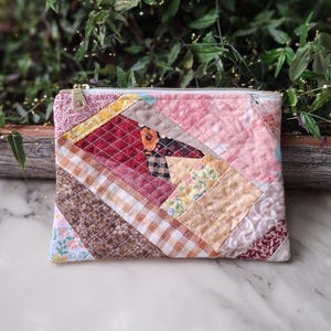 May include: A rectangular patchwork zippered pouch. The pouch features a variety of fabrics in shades of pink, brown, yellow, and red, arranged in a geometric pattern. The zipper is silver-toned, and the background is a marble surface.