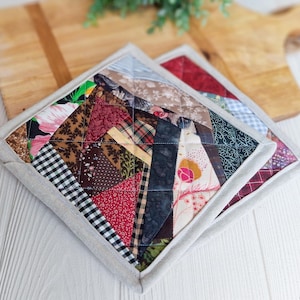 Puede incluir: Dos salvamanteles cuadrados acolchados con un diseño de patchwork. Los salvamanteles presentan una variedad de patrones de tela, incluyendo estampados florales, a cuadros y geométricos, en tonos rojos, marrones y verdes. Cada salvamanteles mide aproximadamente 20 cm cuadrados.