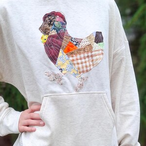 May include: Cream-colored hoodie with a large patchwork chicken design on the front. The chicken is made of various fabric patterns in shades of red, brown, yellow, and blue. Features a kangaroo pocket and a hood.