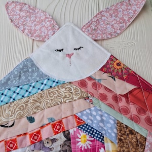May include: A handmade patchwork bunny-shaped quilt with a white face, embroidered eyes and nose, and pink floral ears. The quilt features a variety of colorful fabric patterns, including floral, plaid, and animal prints, stitched together in a chevron design.