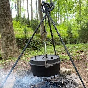 May include: A black cast iron Dutch oven suspended over a campfire. The pot hangs from a black metal tripod. The fire pit is surrounded by rocks and the background shows trees and foliage.