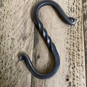 May include: A hand-forged, dark gray metal S-hook. The hook features a twisted central section and rounded ends with small loops. The hook is displayed on a weathered wooden surface, showcasing its rustic aesthetic.