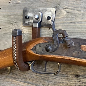Hand-forged brushed steel J-hook wall mount wrapped in brown leather with cross-stitching, riveted to a hammered steel plate and mounted on a rustic wooden wall. Hutton Forge Brushed Steel & Brown Leather gun display hook.